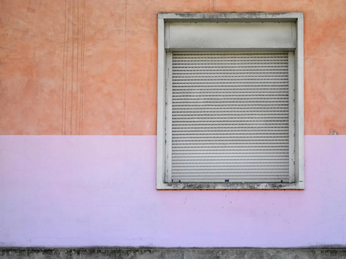 Volet roulant gris clair baissé, coffre apparent, dans un encadrement de fenêtre sur un enduit ocre rose et mauve